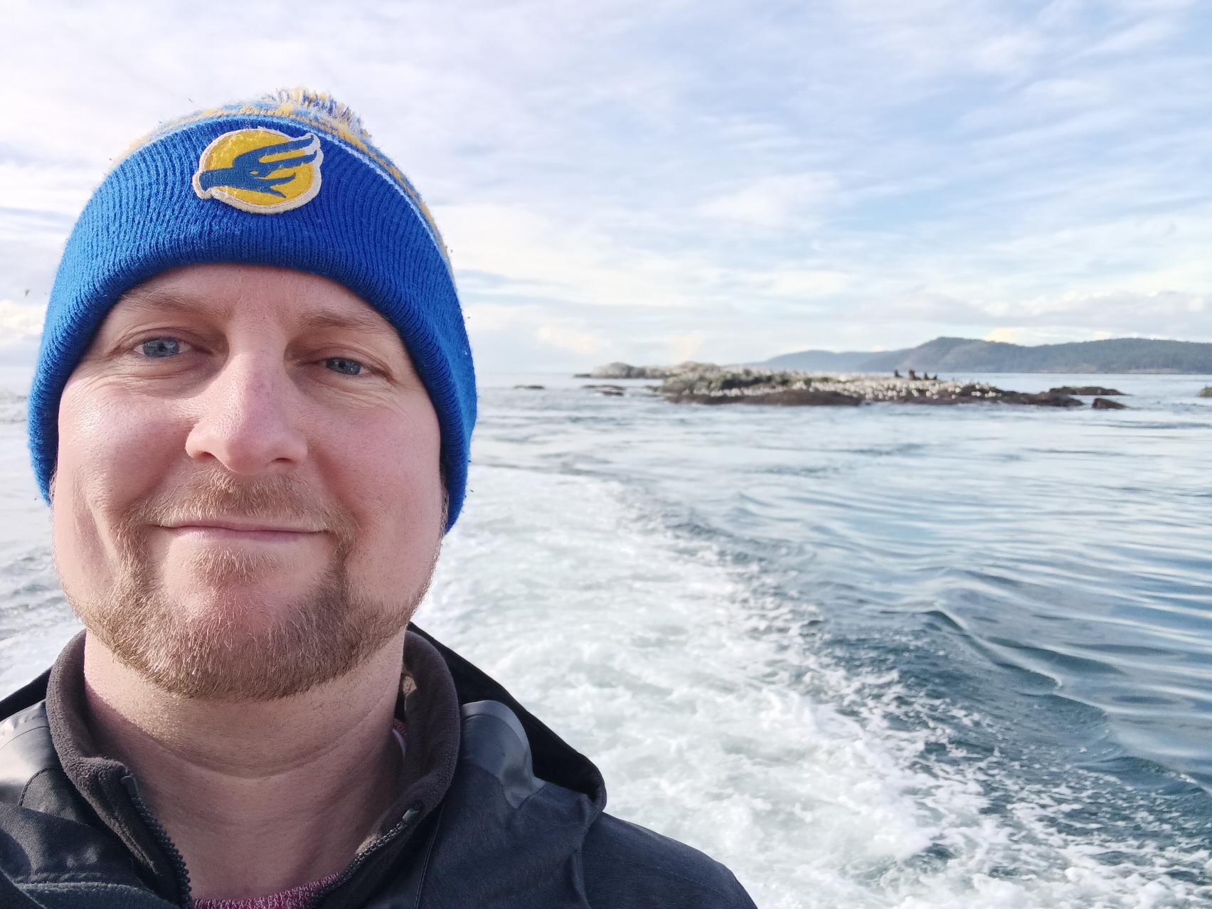 A headshot of Derek Murray, a man wearing a blue toque and black coat, taken on a boat with waves and small islands in the background.